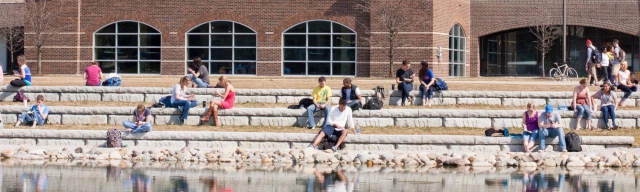 Zumberg pond featuring Kirkhof center in the background and GVSU students sitting near the pond